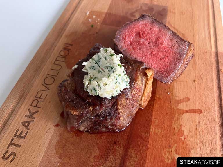 Cooked dry-aged steak topped with compound butter, served on a wooden board.