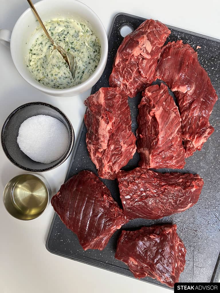 Raw hanger steak pieces on a dark cutting board next to a bowl of garlic herb butter, kosher salt, and cooking oil.