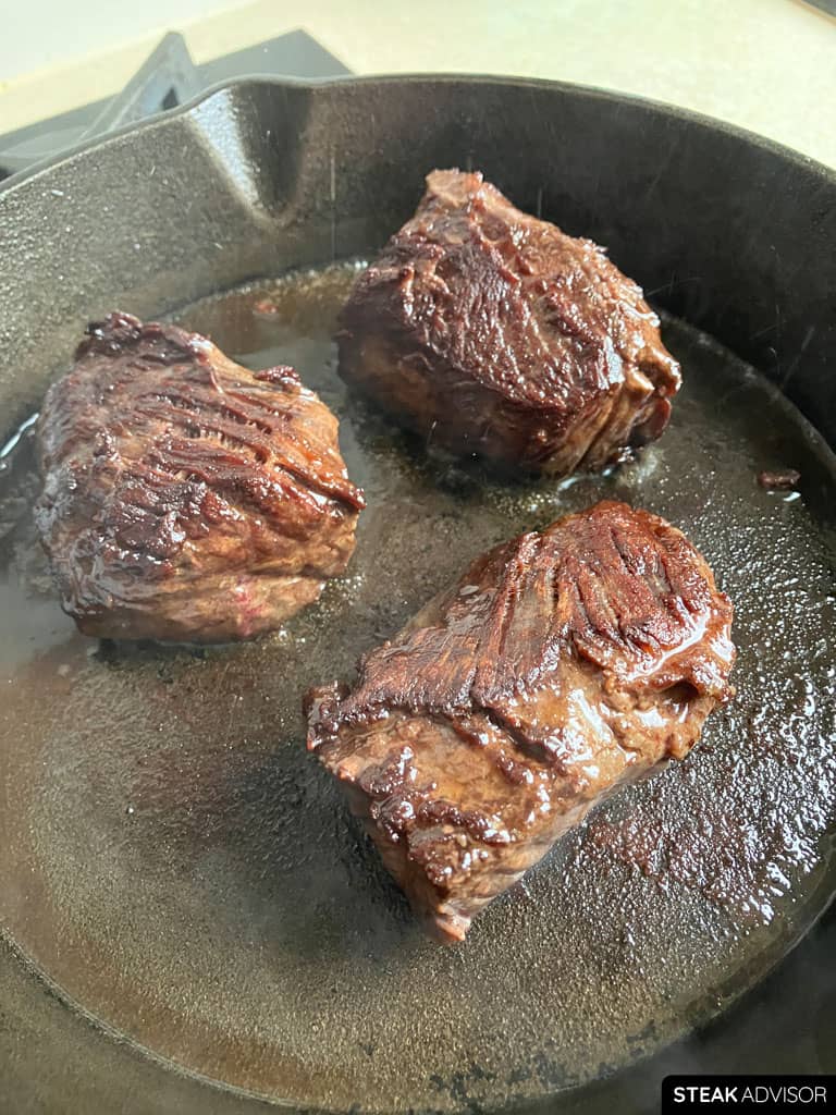 Three hanger steak pieces searing in a hot cast-iron skillet with a deep golden-brown crust.